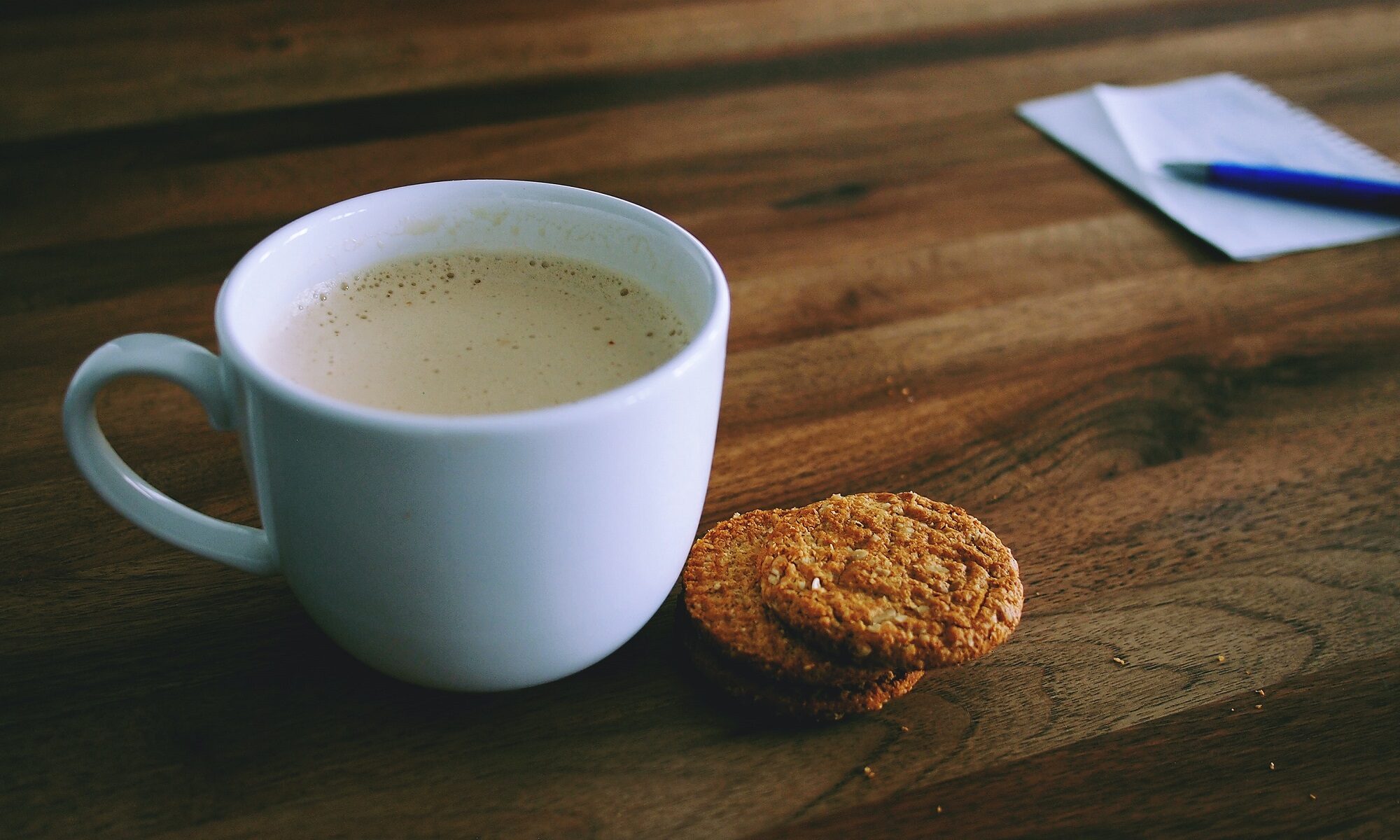 Na zdjęciu małą filiżanką z kawą, obok dwa okrągłe ciastka, w tle kartki z notatnika z długopisem